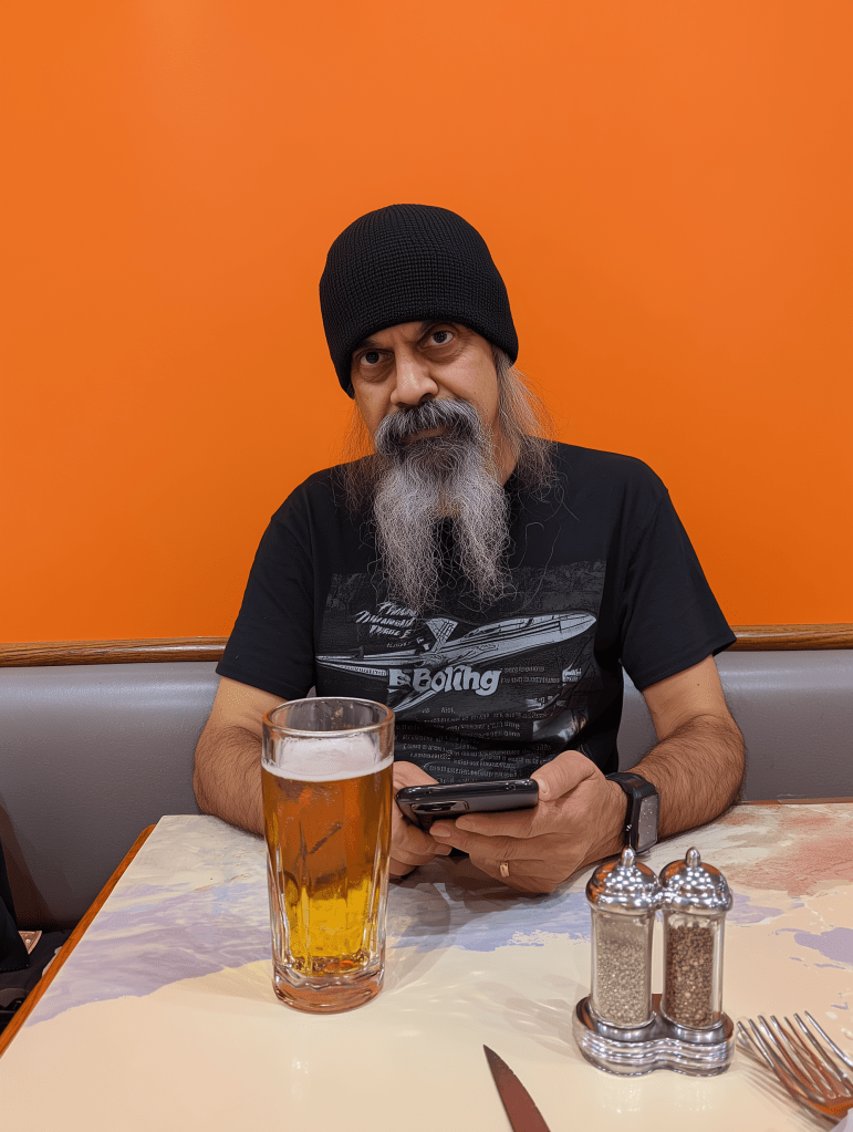 A man with a long beard and wearing a black beanie sits at a table with a large glass of beer, looking at his phone against an orange background.