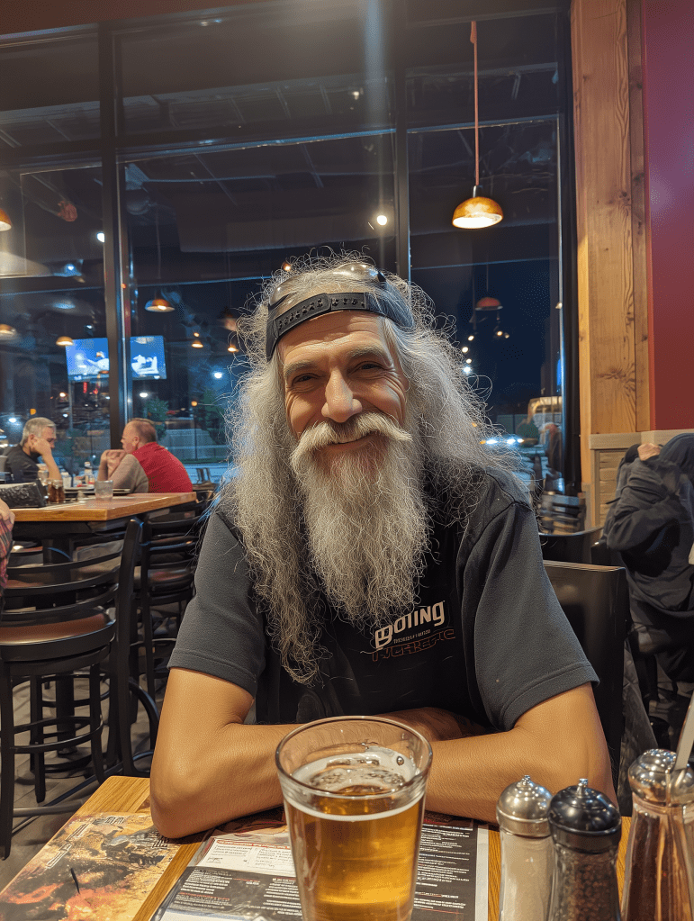 A smiling older man with long gray hair and a beard, wearing a dark t-shirt and a baseball cap, sits at a restaurant table with a glass of beer in front of him.