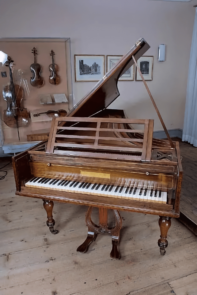 A vintage wooden piano with an open lid and visible keyboard, displayed in a room featuring various string instruments and framed artwork.