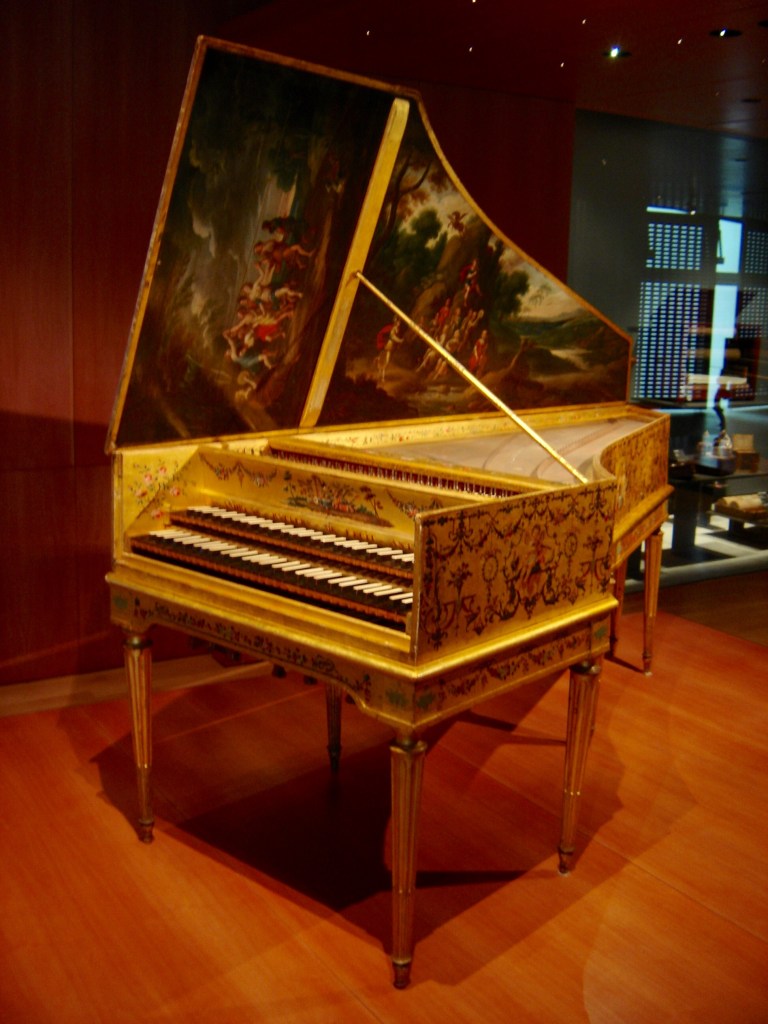An ornate harpsichord with a golden finish and detailed floral designs, featuring a painted lid depicting a pastoral scene.
