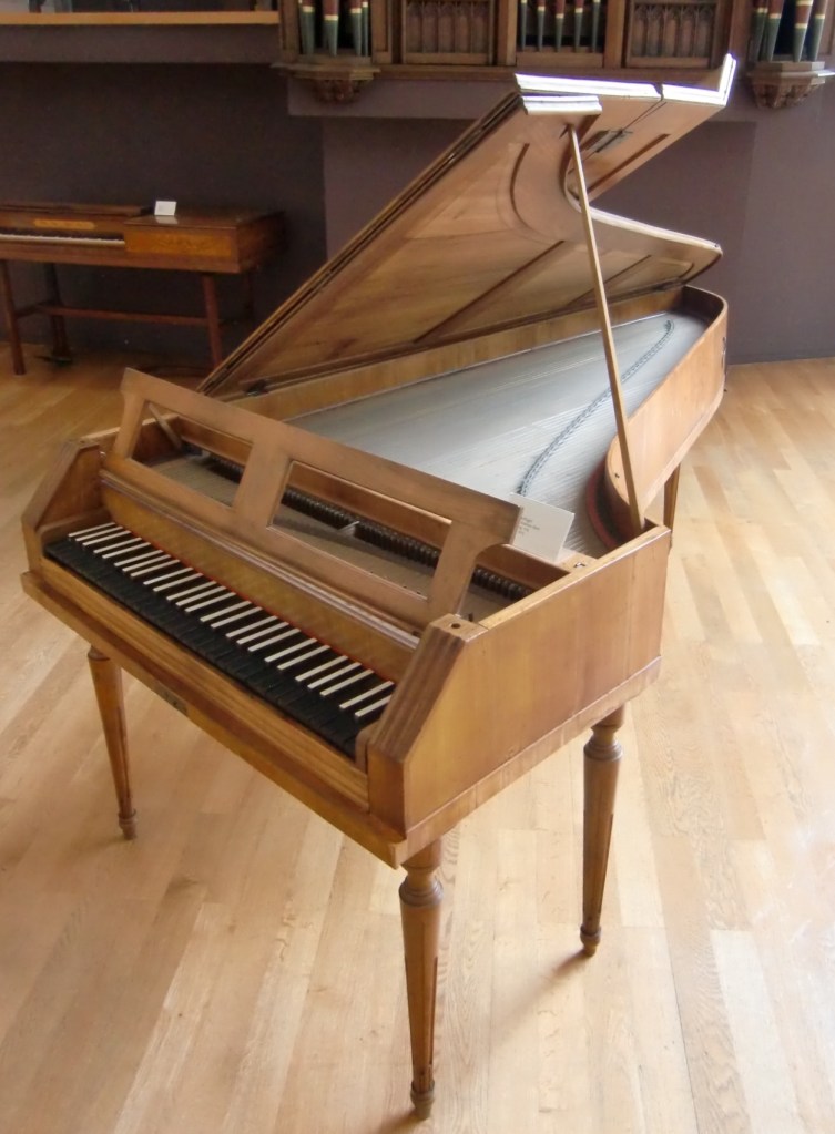 A wooden harpsichord with an open lid, showcasing its keyboard and strings, placed in a well-lit room with wooden flooring.