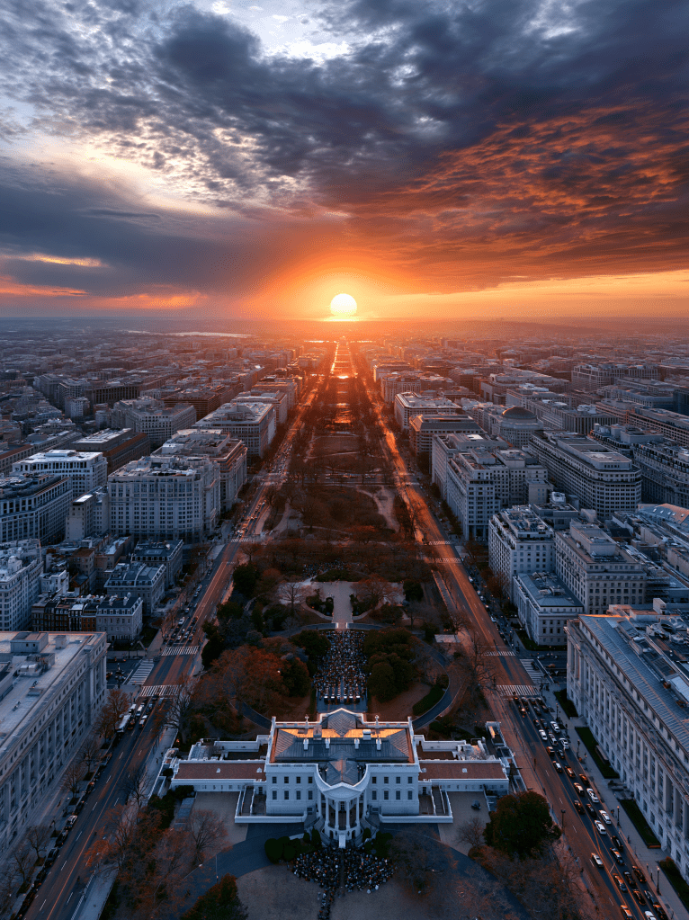 Aerial view of a cityscape at sunset, featuring buildings and streets with a vibrant orange and purple sky.
